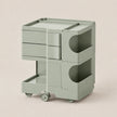 A sage green modular rolling storage cart with three drawers, two open shelves, and caster wheels, shown on a solid light beige background.
