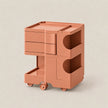 A modern two-tier rolling storage cart in a matte terracotta color, featuring three drawers on the top level, open side compartments, and a bottom pull-out tray on wheels, set against a solid light beige background.