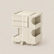 A modern, minimalist two-tier storage cart in a warm off-white/cream color, featuring three drawers on the top tier, open storage compartments with curved cutouts on the side, and a bottom pull-out drawer on wheels, set against a solid light beige background.