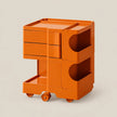 An orange modular plastic storage cart with three drawers, open shelves, and caster wheels, shown on a solid light beige background.