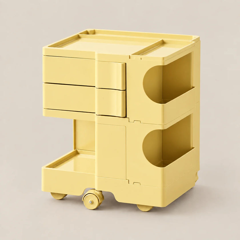 A pale yellow plastic rolling storage cart with three drawers, open side compartments, and caster wheels, shown on a neutral light gray background.