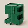 A dark green rolling storage cart with three drawers, open shelving compartments, and caster wheels, shown on a plain beige studio background.