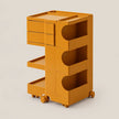 A mustard yellow rolling storage cart with three drawers on the left, three open shelves with curved cutouts on the right, and caster wheels at the base, shown on a plain light beige background.