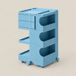 A light blue modular plastic storage cart with three drawers, three open shelves featuring semi-circular cutouts, and four caster wheels, shown on a solid beige background.