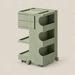 A three-tier rolling storage cart in a muted sage green color, featuring three drawers on the top level, open shelves in the middle, and a lower tray on wheels, set against a solid light beige background.