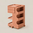 A terracotta-colored plastic rolling storage cart with three drawers, open shelves, and wheels, shown on a solid light beige background.