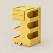 A bright yellow modular rolling storage cart with three drawers, three open shelves, and caster wheels, shown on a clean light beige studio background.