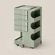 A modern three-tier rolling storage cart in a muted sage green color, featuring three drawers on the left and open storage compartments on the right, set against a solid light beige background.