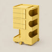A yellow plastic rolling storage cart with three drawers on the left and three open shelves on the right, set against a solid light beige background.