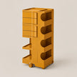 A mustard yellow modular rolling storage cart with drawers, open shelves, and wheels, shown on a plain light beige studio background.