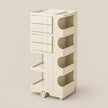 A tall, cream-colored rolling storage cart with multiple drawers, open shelves, and circular cutout details, shown on a plain beige studio background.