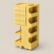 A yellow rolling storage cart with multiple drawers, open shelves, and wheels, shown on a plain light beige studio background.
