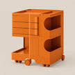 An orange modular plastic storage cart with three drawers on the top left, two open storage compartments with semicircular cutouts on the right, a lower open shelf, and four caster wheels, shown on a light beige background.