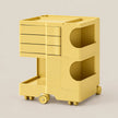 A bright yellow plastic rolling storage cart with three drawers on the top section, two open circular storage compartments, and a lower tray, shown on a light beige background.
