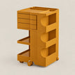 A mustard yellow modular rolling storage cart with three drawers, two open shelves, and wheels, shown on a light beige background.