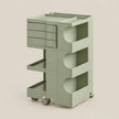 A modern three-tier rolling storage cart in a muted sage green color, featuring three drawers on the top left, two open shelves with circular cutouts, and two caster wheels at the base, set against a solid light beige background.