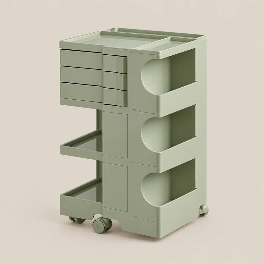 A modern three-tier rolling storage cart in a muted sage green color, featuring three drawers on the top left, two open shelves with circular cutouts, and two caster wheels at the base, set against a solid light beige background.