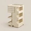 A cream-colored modular plastic storage cart with three tiers, featuring a top drawer unit, open shelves, and caster wheels, set against a solid beige background.
