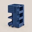 A dark blue modular rolling utility cart with three tiers of shelves and drawers, shown on a solid beige background.
