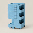 A light blue modular plastic storage cart with three tiers, featuring drawers on the top and two open cubbies on the middle and bottom, set against a solid beige background.