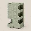 A modern rolling storage cart in a muted sage green color, featuring five drawers on the left, three open storage compartments with circular cutouts on the right, and a lower pull-out tray on casters, set against a solid light beige background.