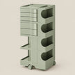A tall, modular storage cart in a muted sage green color, featuring multiple drawers, open shelves, and wheels, set against a solid light beige background.