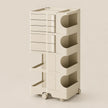 A tall, cream-colored rolling storage cart with multiple drawers, open shelves, and circular cutout compartments, shown on a plain beige studio background.