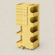 A yellow modular plastic storage cart with four tiers, featuring drawers on the top left, open shelves on the right, and wheels at the base, shown on a neutral beige background.