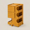 A mustard yellow rolling storage cart with multiple drawers and open shelves, shown on a plain light beige studio background.