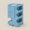 A light blue modular plastic storage cart with three tiers, featuring a stack of drawer-like compartments on the left and open arch-shaped shelves on the right, set against a solid beige background.