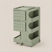 A sage green rolling storage cart with multiple drawers and open shelves, shown on a plain warm beige studio background.