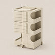 A cream-colored modular storage cart with multiple drawers and open shelves, shown on a clean, solid, warm beige background.