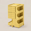 A yellow rolling storage cart with three tiers of drawers and open shelves, shown on a plain light beige studio background.