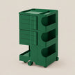 A dark green modular rolling storage cart with multiple drawers and open shelves, shown on a plain beige studio background.