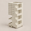 A tall, minimalist rolling storage cart in a warm off-white or cream color, featuring multiple drawers on the left and open shelving compartments on the right, set against a solid neutral background.