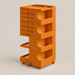 An orange modular plastic storage cart with four tiers, featuring slide-out drawers on the left and open shelves on the right, mounted on wheels against a neutral beige background.