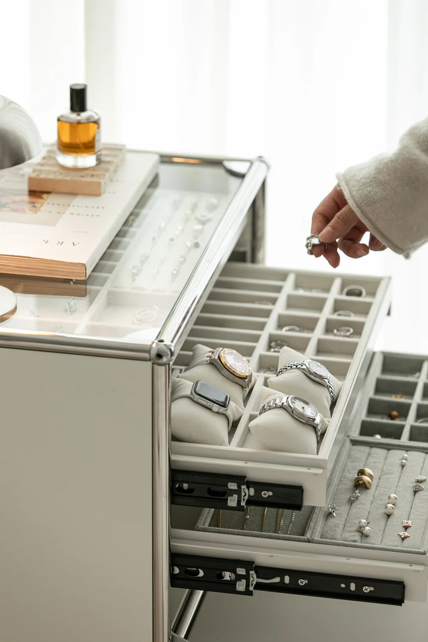 Jewelry drawer with various rings and a hand reaching in to pick one up, with a bottle of perfume on a surface in the background.