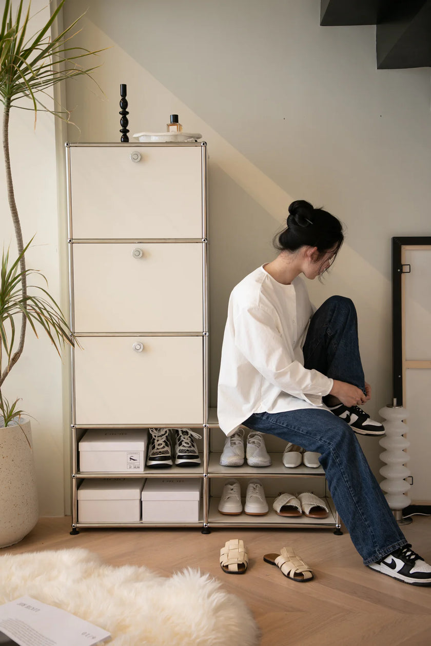 Lady sitting on a shoe rack with shoes around, in a room with a plant and a cabinet.