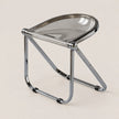 A modern folding stool with a clear smoked-tinted seat and a polished chrome metal frame, shown on a solid light beige background.