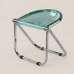 A modern folding stool with a translucent mint green glass seat and a polished chrome metal frame, shown on a solid light beige background.