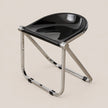 A modern folding stool with a glossy black curved seat and a polished silver metal frame, shown on a solid light beige background.