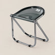 A modern folding stool with a translucent dark gray seat and a polished chrome metal frame, shown on a solid light beige background.