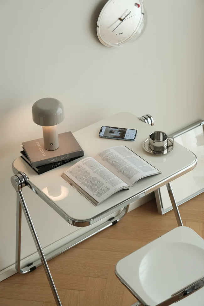 A modern white folding desk with chrome legs, styled with an open book, a gray mushroom-shaped lamp, a smartphone, and a small metal cup on a saucer. The desk is set in a minimalist room with light wood flooring, a cream wall, and a round wall clock.