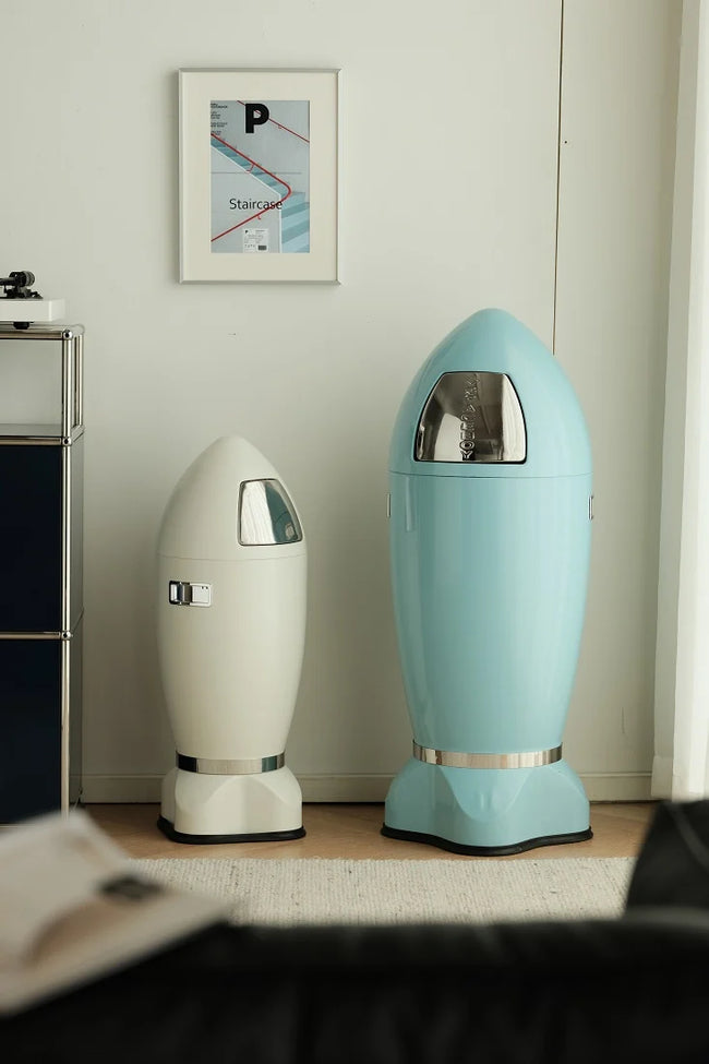 Two retro-futuristic capsule-shaped trash cans standing on a light wood floor against a white wall. One is cream/off-white and the other is light blue, both with metallic silver bands and clear window tops.