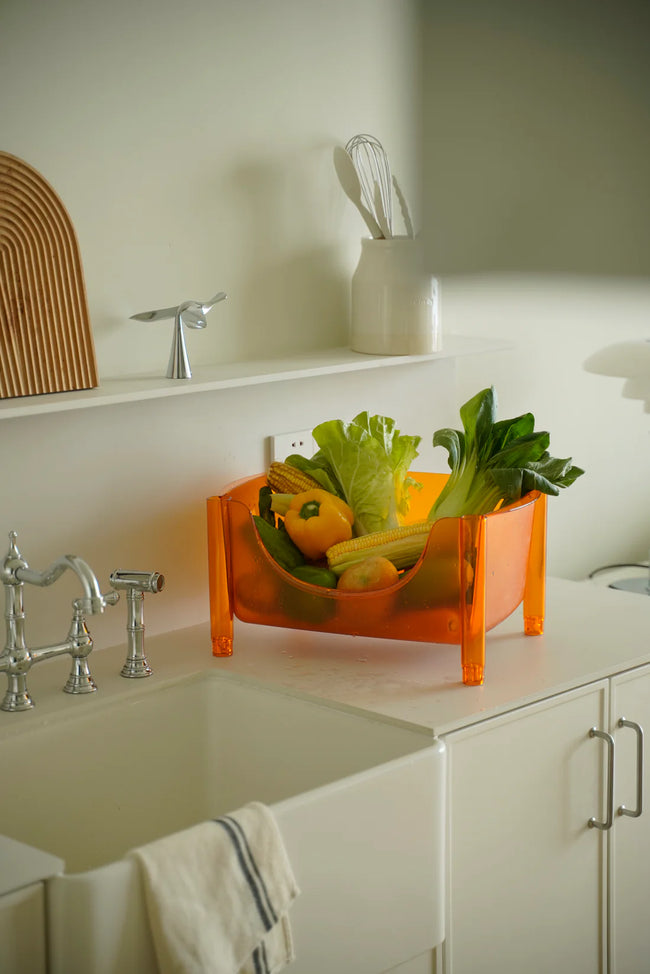 Orange fruit and vegetable plastic storage containers on a kitchen counter with sink and cabinets in the background