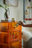 Lady holding a can of beer next to an orange 3 tier rolling cart with bottles on a shelf.