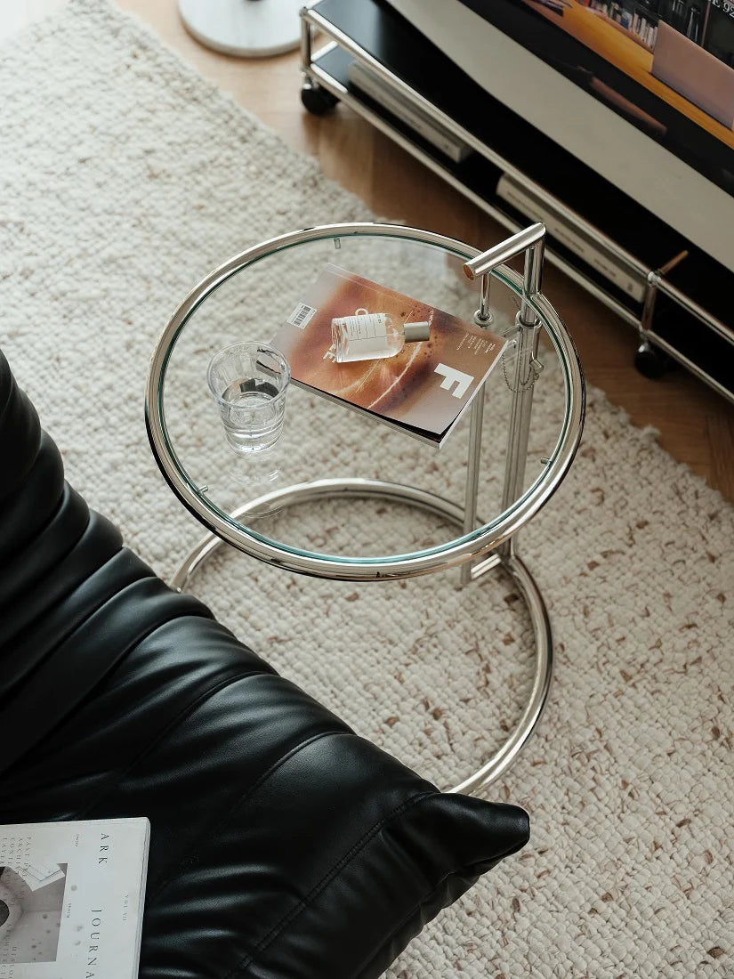 A modern round glass side table with a chrome metal frame, styled in a living room setting next to a black leather chair and a textured cream rug.
