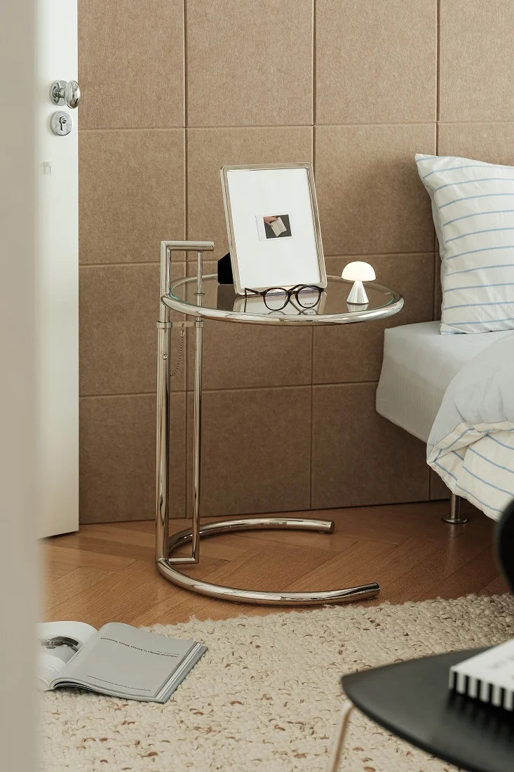 A modern round glass-top side table with a chrome metal frame, styled in a bedroom next to a bed with a striped pillow.
