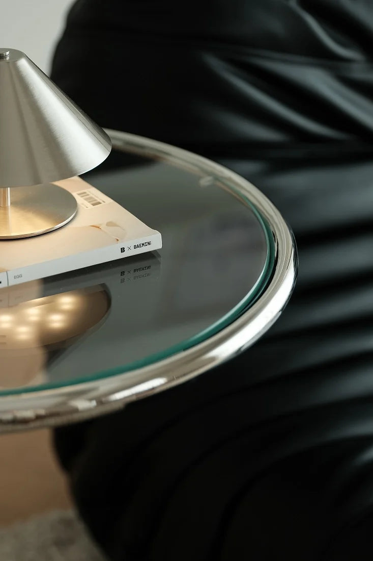 A close-up of a modern round glass side table with a chrome metal rim, holding a silver lamp and a book, next to a person wearing a black leather chair.