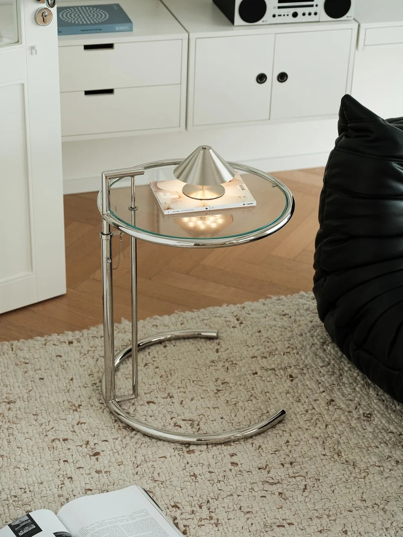 A modern round glass-top side table with a curved chrome metal frame, styled in a living room setting on a beige rug next to a black leather chair.